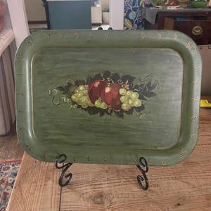 May include: A rectangular olive green metal tray featuring a hand-painted central motif of red apples and green grapes surrounded by dark leaves. The tray has a raised rim decorated with small gold-colored comma-shaped accents along the border.