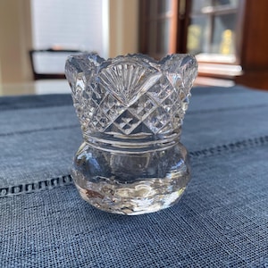 Può includere: Un portacandele in vetro trasparente tagliato con un motivo a diamante e ventaglio. Il piccolo oggetto decorativo in vetro ha una base arrotondata e un bordo smerlato. Il portacandele è appoggiato su una tovaglia blu.