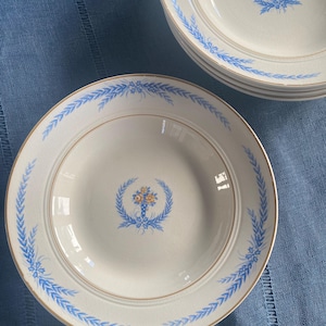 May include: A stack of white ceramic plates with a blue floral design and gold trim. Each plate features a central floral motif and a border of blue leaves. The plates are arranged on a blue surface.