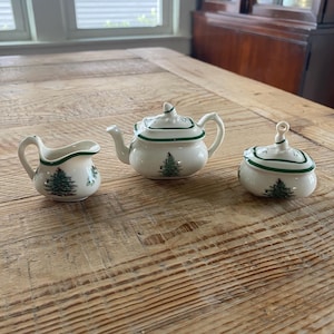 Puede incluir: Un juego de té en miniatura de cerámica que consta de una jarra de leche, una tetera con tapa y un azucarero con tapa. Cada pieza es de color crema con un borde verde oscuro y un gráfico detallado de un pino verde.