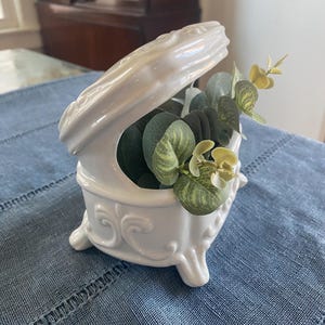 Puede incluir: Una caja de cerámica blanca con tapa abierta, llena de eucalipto artificial verde y amarillo. La caja tiene detalles ornamentados y está sobre un mantel azul. La caja se apoya en pequeños pies.