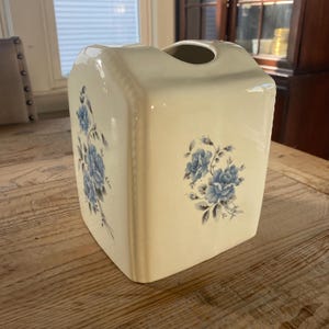 May include: A cream-colored ceramic tissue box cover with a rectangular shape and rounded top edges. The exterior features a delicate blue floral pattern with roses and leaves on the front and side panels, accented by a subtle beaded border detail.