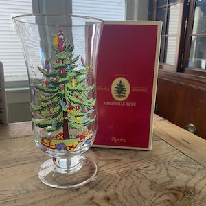 May include: A clear glass vase featuring a colorful Christmas tree graphic with ornaments, a star topper, and wrapped gifts at the base. Beside it stands a red rectangular box labeled "Celebrating Traditions CHRISTMAS TREE Spode".