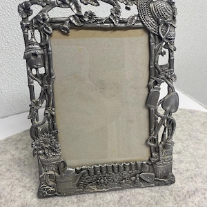 May include: A silver-colored metal picture frame with a garden theme. The frame is decorated with floral and gardening tool motifs, including a watering can, shovel, and flowers. The frame is rectangular and has a neutral-colored backing.