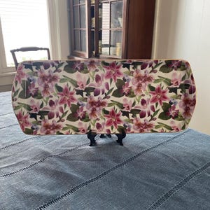 May include: A rectangular serving tray featuring a vibrant floral pattern of pink and purple orchids with green leaves on a white background. The tray has rounded corners and a thin gold-toned rim, displayed upright on a small black stand.