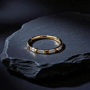 Könnte beinhalten: Ein goldfarbener Bandring mit einer Reihe klarer, rechteckiger Baguette-Steine, die horizontal entlang des äußeren Umfangs eingesetzt sind. Das Metallband hat eine polierte Oberfläche und einen seitlich sichtbaren Klappverschluss.