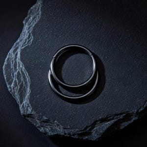 Könnte beinhalten: Ein schwarzer, kreisförmiger Körperschmuck. Der Schmuck besteht aus einem dunklen, glänzenden Metall und besteht aus zwei verbundenen Ringen. Das Stück ist vor einem dunklen, strukturierten Hintergrund platziert.
