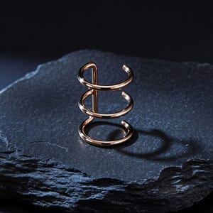 Puede incluir: Un pendiente de oreja de oro rosa con tres bandas curvas apiladas, conectadas por una barra vertical. La joya se encuentra sobre una superficie oscura y texturizada, proyectando una sombra. El diseño es minimalista y moderno.