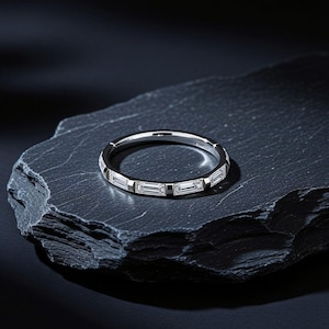 Könnte beinhalten: Ein silberfarbener Bandring mit einer Reihe klarer, rechteckiger Baguette-Schliff-Steine, die horizontal um den gesamten Ringumfang angeordnet sind. Das Metall hat eine polierte, reflektierende Oberfläche und die Steine sind gleichmäßig verteilt.