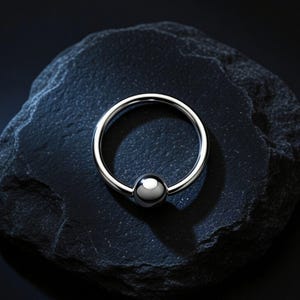 May include: A circular silver-toned captive bead ring featuring a single polished spherical ball closure. The metal hoop has a smooth, reflective surface and a consistent gauge throughout its circumference, designed for body piercing applications.
