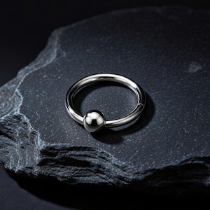 Puede incluir: Un anillo circular de color plateado con un cierre de bola esférica pulida. El aro de metal tiene una superficie lisa y reflectante con un diseño sin costuras, mostrando una forma circular clásica con un componente de bola central.