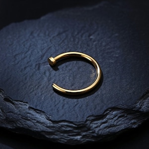 May include: A gold-toned circular nose ring featuring a smooth, polished finish and a small flat disc at one end of the open hoop design. The jewellery piece is crafted with a simple, continuous curved wire that forms a partial circle shape.