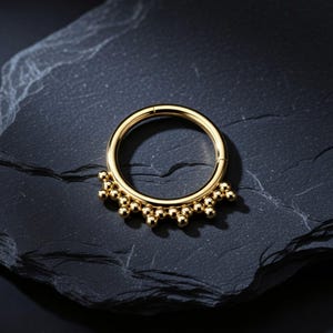 Op de afbeelding: Een cirkelvormige goudkleurige ring met een reeks kleine, uniforme gouden bolletjes bevestigd langs de onderste helft van de buitenrand. Het sieraad heeft een gladde, gepolijste metalen afwerking en een scharnierend segment om te openen.