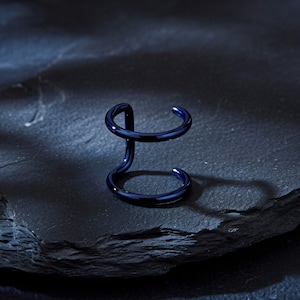 May include: A dark blue ear cuff with a double-loop design. The jewelry piece is made of a shiny, smooth material and sits on a dark, textured surface. The ear cuff has a modern, minimalist aesthetic.