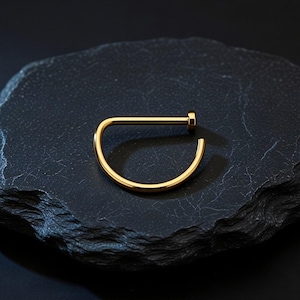 Könnte beinhalten: Ein goldfarbener Nasenring in D-Form. Der Schmuck besteht aus glänzendem, glattem Metall und ist vor einem dunklen, strukturierten Hintergrund platziert. Der Nasenring hat einen geraden Pfosten und eine gebogene, offene Schlaufe.
