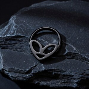 May include: A black nose ring with a silver alien face design. The ring is circular with a hinged closure. The alien face is a stylized shape with two eye-like openings and a curved mouth.