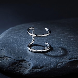 May include: A silver-colored ear cuff with a unique double-band design. The cuff curves around the ear with two circular bands connected by a vertical bar. The jewelry is displayed on a dark gray stone surface.