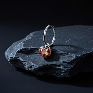 Peut inclure: Boucle d'oreille argentée avec un pendentif en forme de cœur en pierre précieuse orange. La pierre précieuse est facettée et sertie dans un cadre argenté. La boucle d'oreille est présentée sur une surface de pierre texturée gris foncé.
