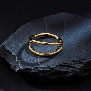 Pode incluir: Um anel dourado com um design de banda dupla. O anel é feito de um metal brilhante e reflexivo e repousa sobre uma superfície texturizada cinza escuro. O design do anel apresenta duas bandas curvas paralelas que se cruzam.