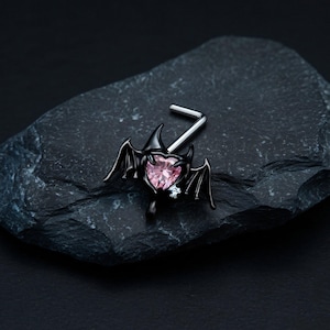 Puede incluir: Un piercing de nariz en forma de L con un engaste de metal negro con forma de alas de murciélago que rodea una piedra preciosa rosa en forma de corazón. Una pequeña piedra redonda y transparente se sitúa en la base del ala derecha.