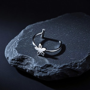 Può includere: Un anello per septum in metallo color argento con un piccolo ciondolo a forma di fiore a cinque petali fissato alla banda circolare. Il gioiello presenta una finitura liscia e lucida con un perno dritto sulla parte superiore per il piercing.