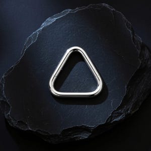 Può includere: Un gioiello triangolare argentato e lucido. Il design geometrico risalta su uno sfondo scuro e strutturato, evidenziando il metallo lucido. Il gioiello sembra essere un ciondolo o un orecchino.