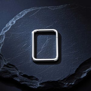 May include: A silver-colored, rectangular clasp, possibly for a bracelet or necklace, is displayed on a dark, textured surface. The clasp is a simple, geometric shape with a hinged closure, reflecting light.