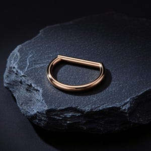 Puede incluir: Un anillo de piercing en forma de D en oro rosa. La joya es de metal brillante y está colocada sobre un fondo oscuro y texturizado. El anillo es un semicírculo con una barra recta en la parte superior.