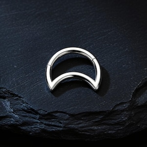 Puede incluir: Un anillo de segmento articulado de color plateado con forma de media luna. La pieza de joyería tiene un acabado metálico liso y pulido, con una banda circular que se transforma en un diseño curvo y puntiagudo en la base, creando una silueta lunar.