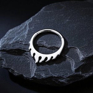 May include: A silver-toned circular hinged ring featuring a decorative flame-shaped design along the bottom half. The metal surface is polished and reflective, showcasing a smooth finish with distinct, curved cutouts forming the flame pattern.