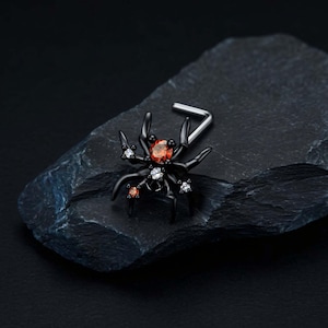 Pode incluir: Um piercing de nariz preto em forma de L com um design de aranha. O corpo da aranha é acentuado com uma pedra preciosa laranja central, rodeada por pedras mais pequenas transparentes e laranjas incrustadas nas pernas e na estrutura de metal.