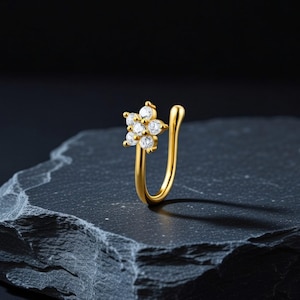 May include: A gold-colored nose ring featuring a flower design with six clear, round-cut gemstones. The ring has a curved, U-shaped design for easy wear. The jewelry is displayed on a dark, textured surface.
