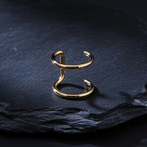 Könnte beinhalten: Ein goldfarbener Ear Cuff mit einem Doppelring-Design und einer glatten, polierten Oberfläche. Das Schmuckstück besteht aus zwei parallelen, kreisförmigen Bändern, die durch einen vertikalen Steg verbunden sind und eine offene Struktur bilden.