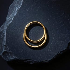 Pode incluir: Um anel de segmento articulado em tom dourado com um design circular liso e um elemento curvo secundário fixado na parte inferior do aro, criando uma aparência em camadas com um acabamento metálico polido e reflexivo.