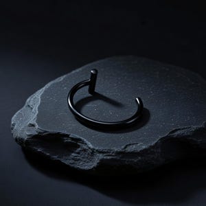 May include: A matte black metal cuff bracelet featuring a circular band with a single vertical cylindrical post extending upward from one end. The design is open-ended, showcasing a smooth, dark finish that contrasts with the textured surface.