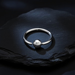 May include: A silver-colored captive bead ring with a polished, spherical bead. The ring is circular with a small gap for insertion. The jewelry is displayed on a dark, textured surface.