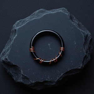 May include: A circular black hoop ring featuring a thin copper wire wrapped around the lower portion of the band. The ring has a smooth, polished finish and a hinged closure mechanism, creating a contrast between the dark metal and copper.