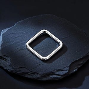 Könnte beinhalten: Ein polierter, quadratischer Klapp-Creolenring in Silberoptik mit einer glatten, reflektierenden Metalloberfläche. Das Schmuckstück verfügt über einen nahtlosen Verschlussmechanismus und abgerundete Ecken für ein klares, geometrisches Design.