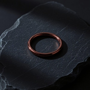 Può includere: Un anello circolare con una finitura liscia e lucida color rame scuro. L'anello presenta un design a segmento incernierato senza cuciture, che consente di aprirlo e chiuderlo in modo sicuro. La superficie metallica riflette la luce.