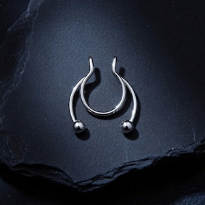 Può includere: Un anello per setto color argento caratterizzato da un design a cerchio con due piccole perle sferiche alle estremità. Il gioiello presenta una finitura metallica liscia e lucida e una struttura aperta e curva che forma una figura simmetrica.