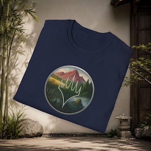 May include: A navy blue t-shirt featuring a circular graphic on the chest. The graphic depicts a landscape with a red mountain, green trees, and a body of water, overlaid with a white, five-pointed, crown-like symbol enclosed in a silver border.