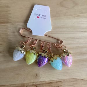 May include: Six strawberry-shaped stitch markers in various pastel colours, including white, yellow, purple, blue, and pink. Each marker has a gold-coloured stem and is attached to a copper safety pin. A tag reads "Handmade with Love".