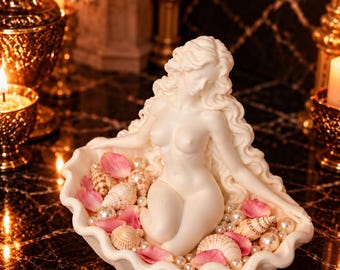 Peut inclure: Une sculpture blanche représentant une silhouette aux longs cheveux flottants assise dans un grand coquillage ouvert. La coquille est remplie de petites perles blanches, de divers coquillages en spirale et de pétales de fleurs roses.
