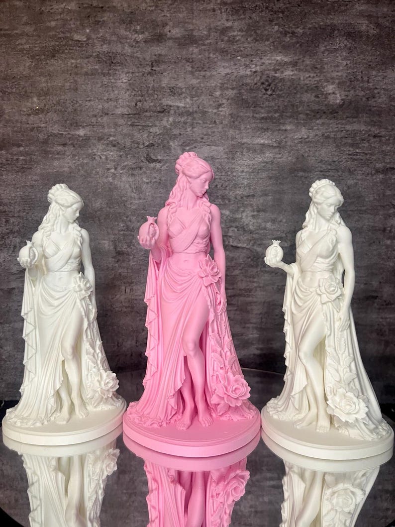 Three Persephone statues displayed together, featuring two elegant white Greek goddess sculptures and one pink centerpiece. Inspired by Greek mythology, Persephone, Queen of the Underworld, holds a symbolic pomegranate while wearing flowing drapery decorated with detailed roses. This comparison photo showcases available color options and handcrafted 3D printed details, perfect for altar decor, pagan decor, mythology collections, witchcraft spaces, and feminine home decor.
