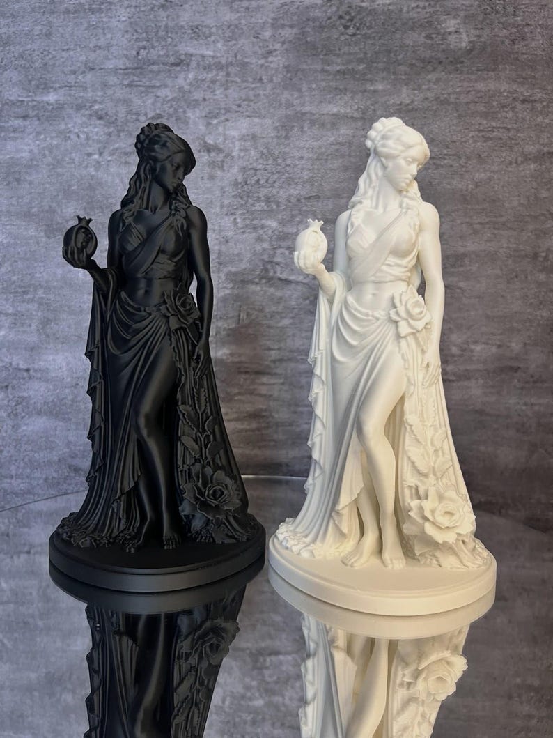 Black and white Persephone Statue displayed side by side to compare available color options. This detailed Greek Goddess sculpture features a symbolic pomegranate, flowing classical robes, and floral accents inspired by Greek mythology. Perfect for pagan altar decor, witchcraft decor, mythology collections, gothic home decor, and lovers of ancient Greek goddesses.