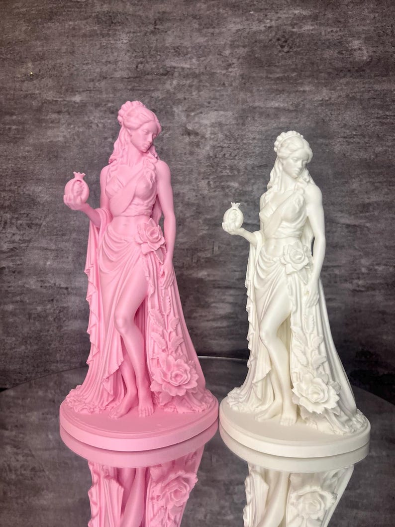 Two Persephone statues shown side by side in pink and white PLA, highlighting the elegant design inspired by Greek mythology. The Goddess of Spring and Queen of the Underworld is depicted holding a symbolic pomegranate with flowing robes and blooming roses, representing rebirth, abundance, and transformation. This comparison image displays available color choices for mythology lovers, pagan altar decor, witchcraft decor, spiritual gifts, and aesthetic home decoration.