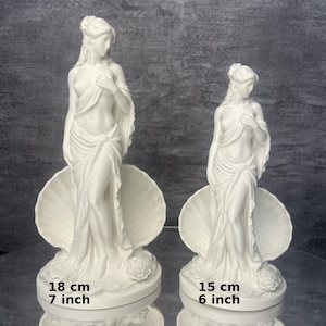 Op de afbeelding: Twee witte beelden van een staande figuur gedrapeerd in stof, geplaatst voor een grote schelp. Het grotere beeld meet 18 cm en het kleinere beeld meet 15 cm. Beide figuren zijn voorzien van een roos aan de basis van het beeldhouwwerk.