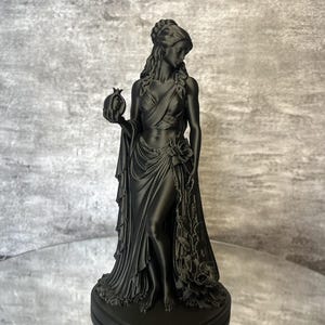 Matte black Persephone Statue displayed in natural daylight, featuring the Greek Goddess holding a detailed pomegranate and wearing flowing robes decorated with elegant roses. This handcrafted Greek mythology sculpture is ideal for pagan altar decor, witchcraft decor, gothic home decor, mythology collections, spiritual spaces, and unique fantasy-inspired gifts.