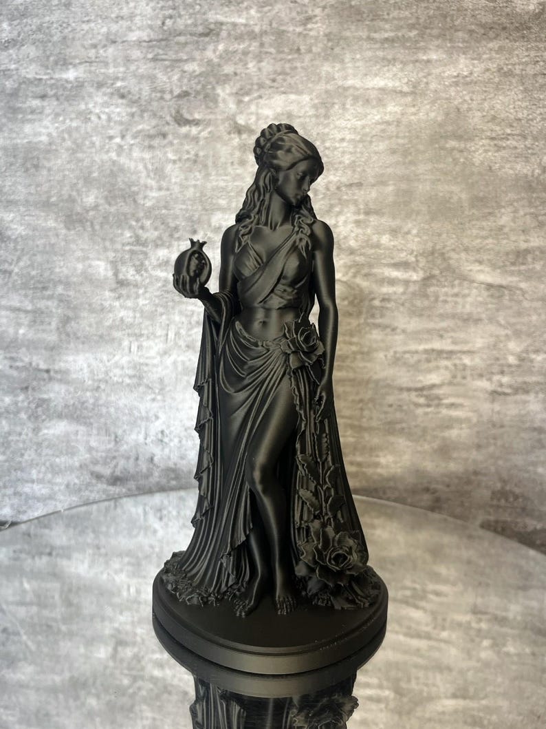 Matte black Persephone Statue displayed in natural daylight, featuring the Greek Goddess holding a detailed pomegranate and wearing flowing robes decorated with elegant roses. This handcrafted Greek mythology sculpture is ideal for pagan altar decor, witchcraft decor, gothic home decor, mythology collections, spiritual spaces, and unique fantasy-inspired gifts.
