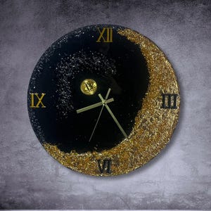 May include: A round wall clock with a black and gold design. The clock face is divided into two sections: black with glitter and gold with glitter. Gold Roman numerals mark the hours. The clock hands and a small coin in the center are gold.